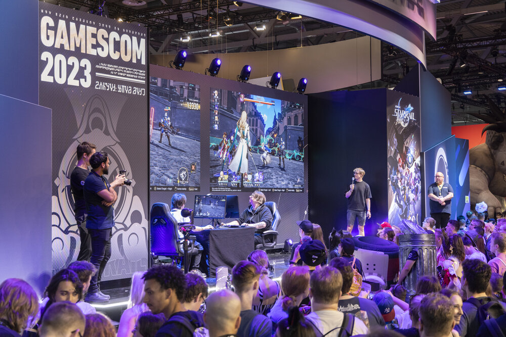 A stage at Gamescom 2023 where two people are playing a computer game, which is being broadcast on a screen in front of an audience.