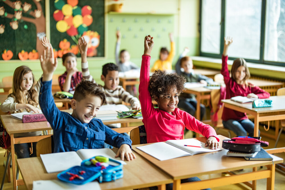 Schoolchildren are sitting at their desks in a classroom; several are raising their hands and laughing.