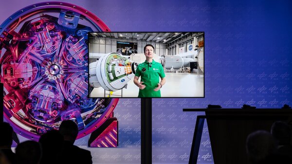 A vibrant control room with colorful machinery is depicted. A screen displays a person in a green shirt explaining in a lab setting, creating a futuristic, informative atmosphere.