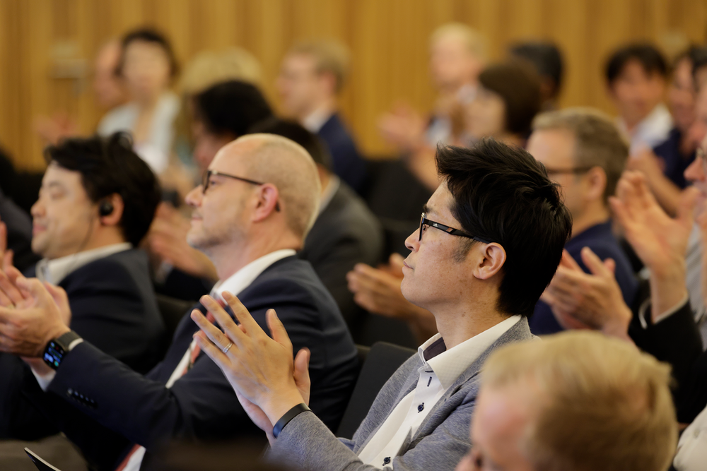 People in business attire are sitting and applauding at a conference in a warmly lit room. The mood is positive and engaged, indicating approval.