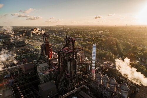 Luftaufnahme eines Industrielandschaft bei Sonnenuntergang, mit rauchenden Schornsteinen und umliegender Landschaft.