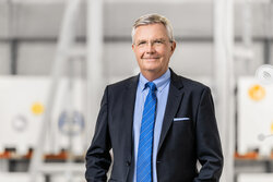 Ein Mann im Anzug mit blauer Krawatte und Brille steht vor unscharfem industriellen Hintergrund und blickt in die Kamera.