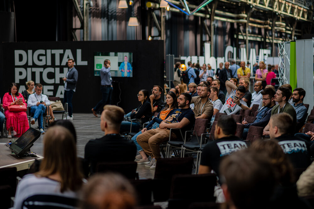 Many people are sitting in the “Digital Pitch Area” of an event hall, watching a presentation on stage.