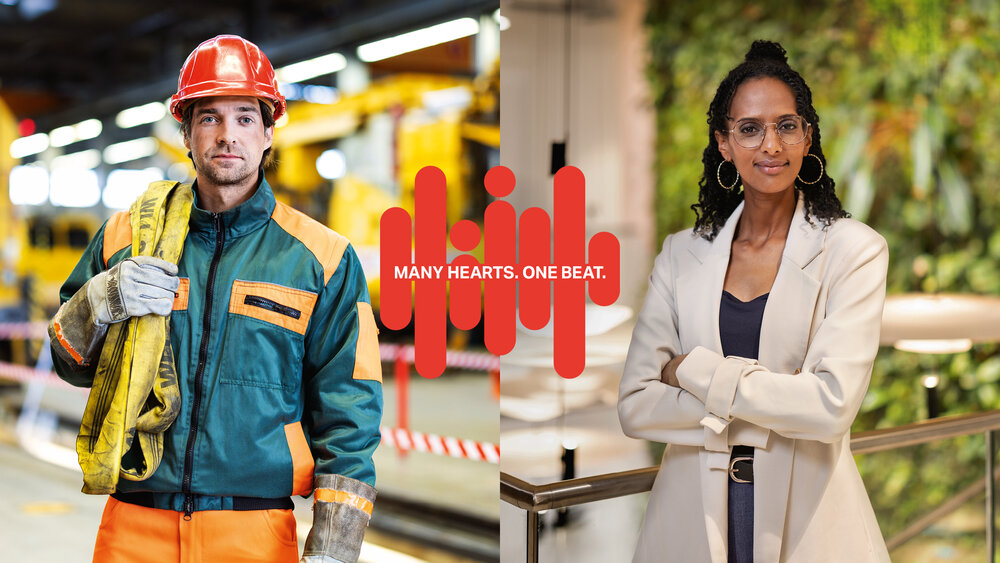 Split image: Left shows a male construction worker in safety gear in a workshop; right shows a female professional in formal attire, arms crossed, in a modern office with greenery. Center text reads "Many Hearts. One Beat." The tone is collaborative and diverse.