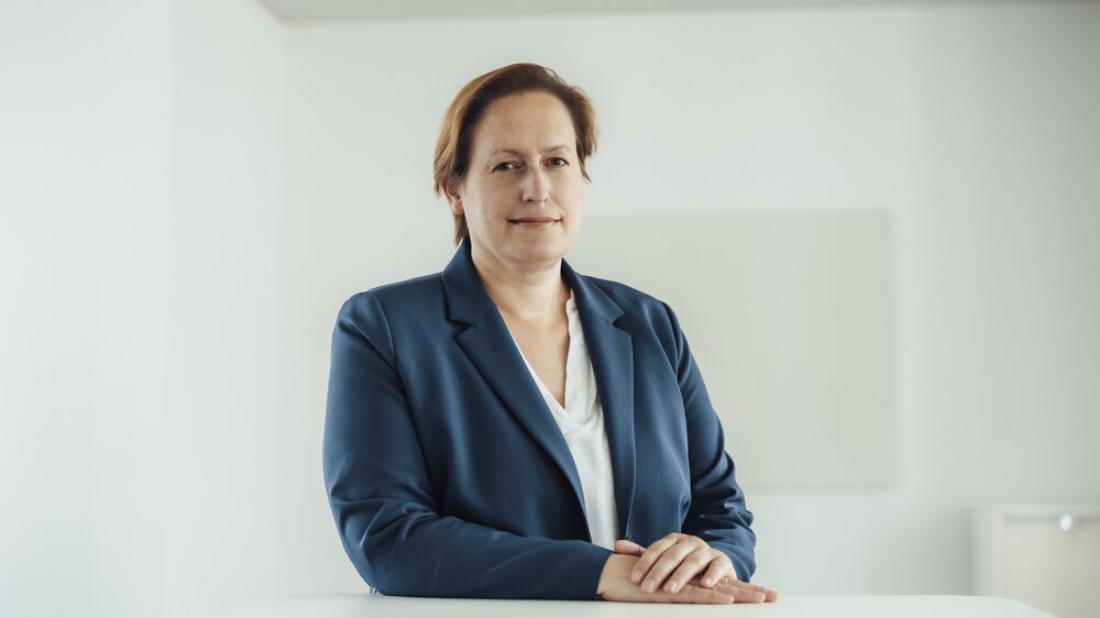 A portrait photo of a smartly dressed woman in a velvet blue blazer. She is sitting behind an office desk with her hands resting on it.