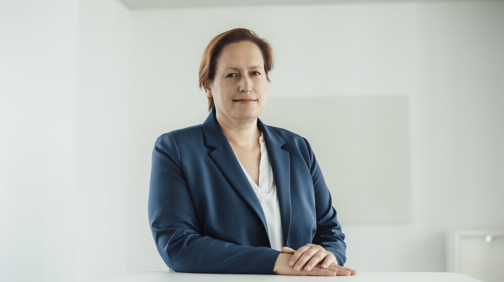 A portrait photo of a smartly dressed woman in a velvet blue blazer. She is sitting behind an office desk with her hands resting on it.