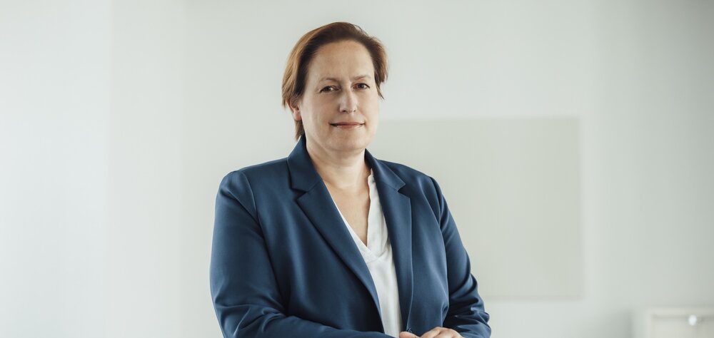 A portrait photo of a smartly dressed woman in a velvet blue blazer. She is sitting behind an office desk with her hands resting on it.