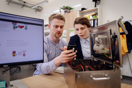 Zwei Personen sitzen vor einem offenen technischen Gerät und schauen auf das Display eines Smartphones.
