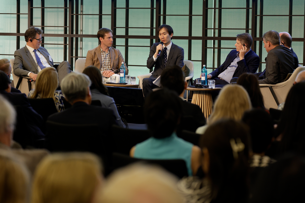 A panel of six men in business attire sits on stage, engaged in discussion. One man holds a microphone. Audience members attentively listen.