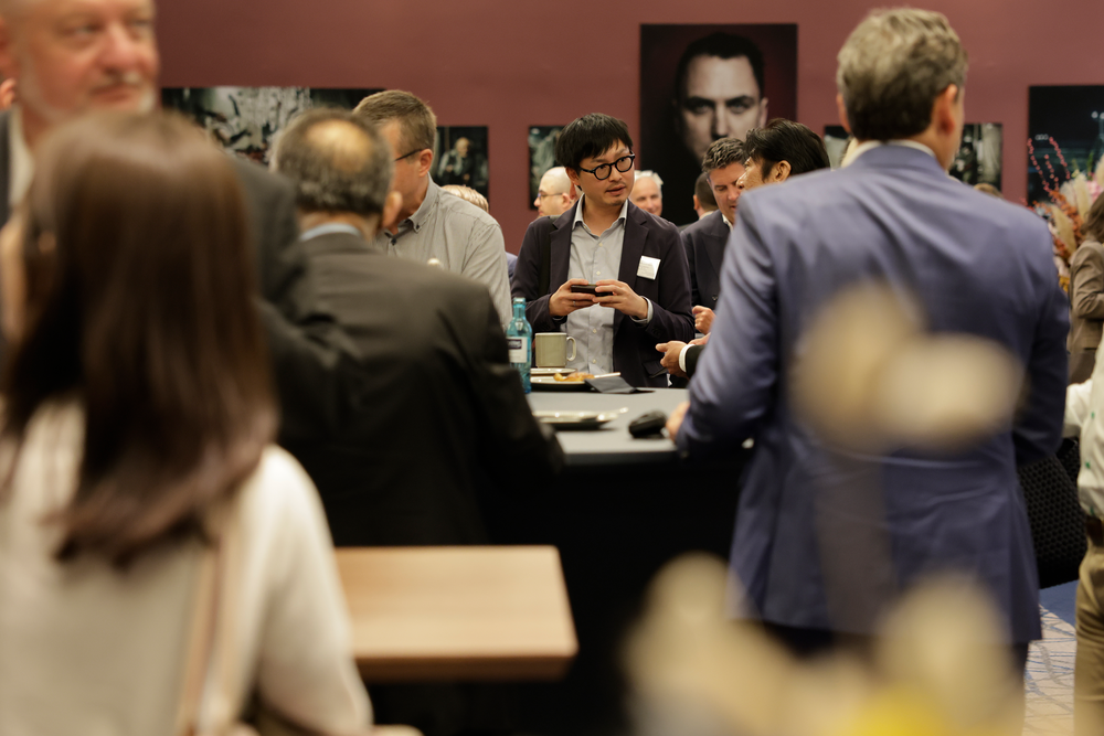A group of people in business attire gathers around a table at a networking event. One person focuses on their phone, surrounded by lively conversation.