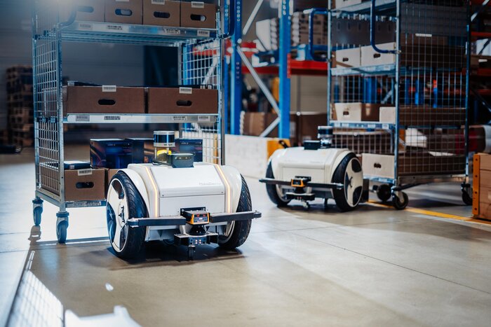 Zwei autonome Transportroboter auf Rädern bewegen Gitterwagen mit Kartons in einem Lagerhaus.