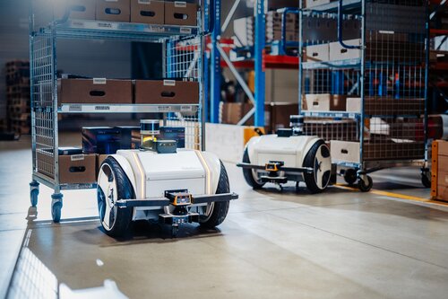 Zwei autonome Transportroboter auf Rädern bewegen Gitterwagen mit Kartons in einem Lagerhaus.
