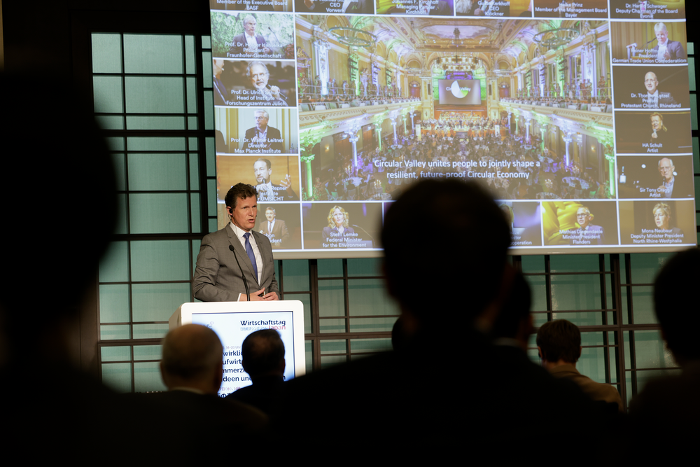 A speaker presents at a podium in front of a slideshow. The screen displays portraits of professionals and a caption about a circular economy. Audience members are silhouetted in the foreground.