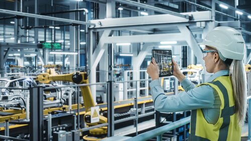 Eine Frau mit Schutzhelm und Warnweste steht in einer Autofabrikhalle und erfasst Roboterarme per Kamera eines Tablets.