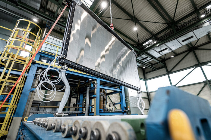 Eine große metallische Baukomponente wird in einer modernen Industriehalle per Kran und Gestell montiert.