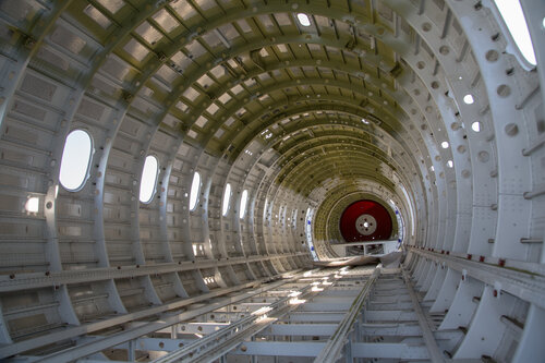 Interior view of an airplane fuselage under construction, with circular ribbing and oval windows. The space is industrial and symmetrical, evoking a sense of engineering precision.