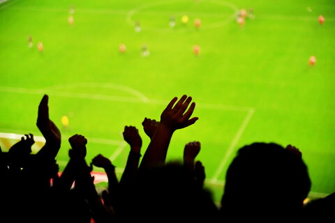 Eine Silhouette jubelnder Fußballfans im Stadion mit erhobenen Armen. Im Hintergrund ist ein verschwommenes Spielfeld mit Spielern zu sehen.