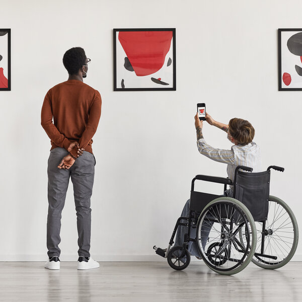 Zwei Personen betrachten Kunstbilder in einer Galerie. Eine Person steht, die andere sitzt im Rollstuhl und fotografiert ein Bild mit dem Smartphone.