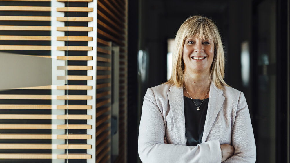 A portrait photograph of a smartly dressed woman. She has her arms crossed and is wearing a light grey blazer and a necklace.
