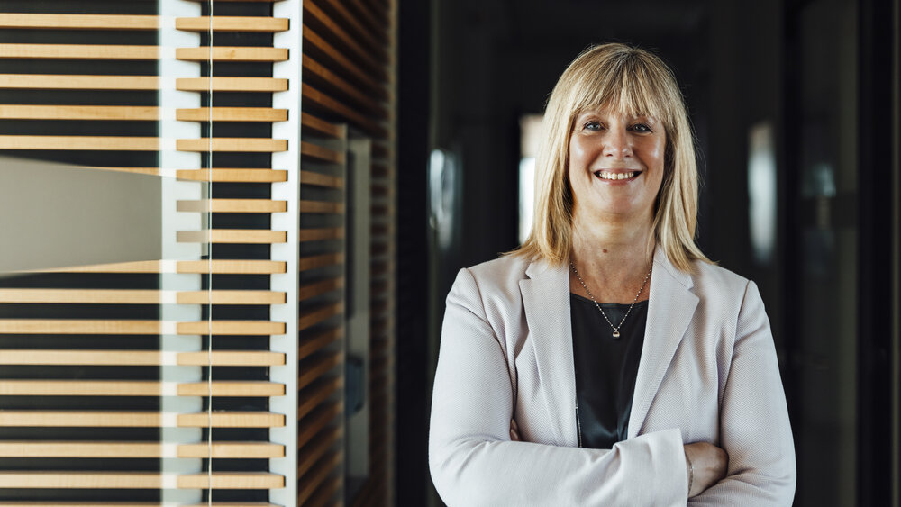A portrait photograph of a smartly dressed woman. She has her arms crossed and is wearing a light grey blazer and a necklace.
