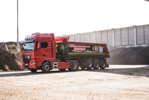 Ein roter Lkw mit einem Anhänger für Schüttgut, beschriftet mit Schüttflix, steht auf einem Baustoffhof vor mehreren Erdhaufen.