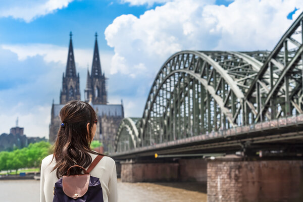 Eine Frau mit Rucksack steht am Rheinufer und blickt auf die Hohenzollernbrücke und den Kölner Dom.