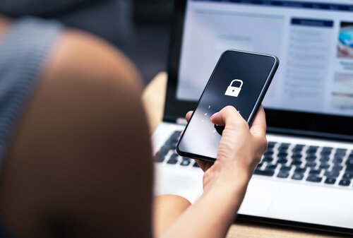Person hält ein Smartphone mit angezeigtem Schloss-Symbol zur Entsperrung vor einem geöffneten Laptop.