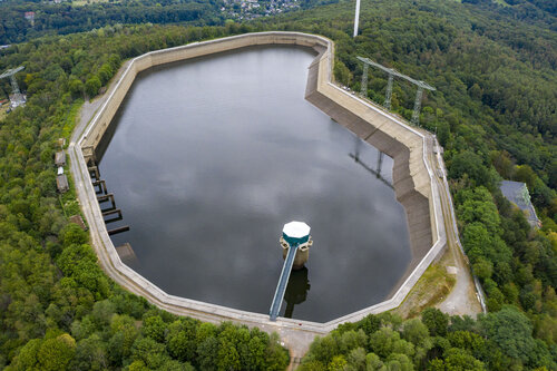 Eine Luftaufnahme eines großen Wasserbeckens, umgeben von Betonwänden, eingebettet in einen dichten Wald mit Strommasten am Rand.
