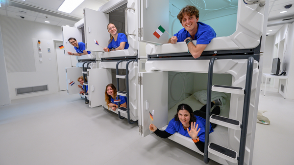 Six people in blue shirts happily lean out of futuristic sleep pods stacked in two rows. Each holds a small flag, creating a cheerful and playful atmosphere.