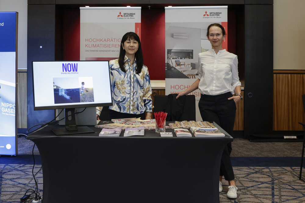 Two women are standing behind an exhibition stand with brochures and a monitor, with two Mitsubishi Electric company banners in the background.