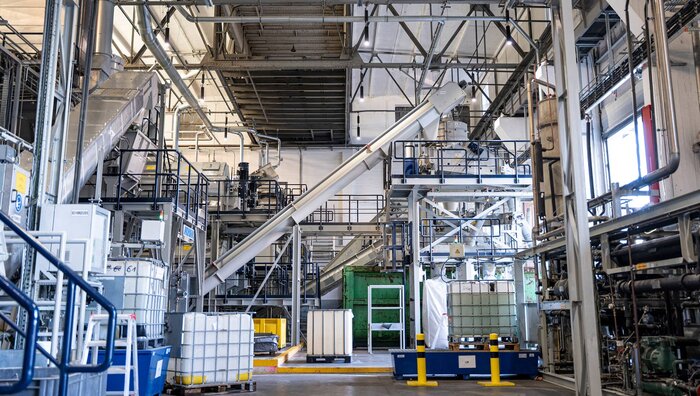 Eine industrielle Produktionshalle mit Förderanlagen, Tanks und Rohrleitungen, im Vordergrund mehrere weiße IBC-Container.