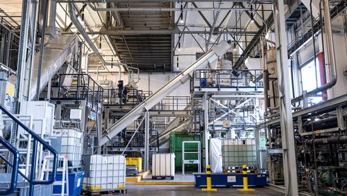 Eine industrielle Produktionshalle mit Förderanlagen, Tanks und Rohrleitungen, im Vordergrund mehrere weiße IBC-Container.