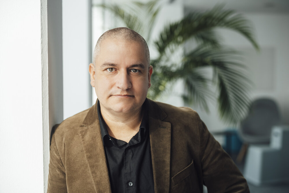 A portrait photograph of a man wearing a brown jacket and black shirt standing indoors with a plant and furniture in the background.