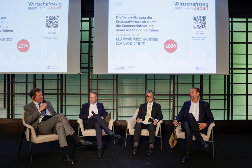 Four speakers sit on a panel with a backdrop displaying "Wirtschaftstag Japan 2026." They are engaged in discussion, conveying a formal tone.