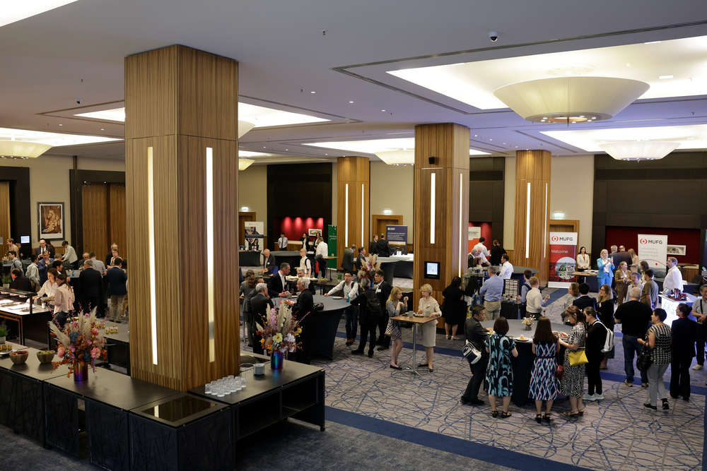 Spacious conference room with groups of people networking. Tall wooden pillars divide the space. Tables with colorful flower arrangements are scattered.