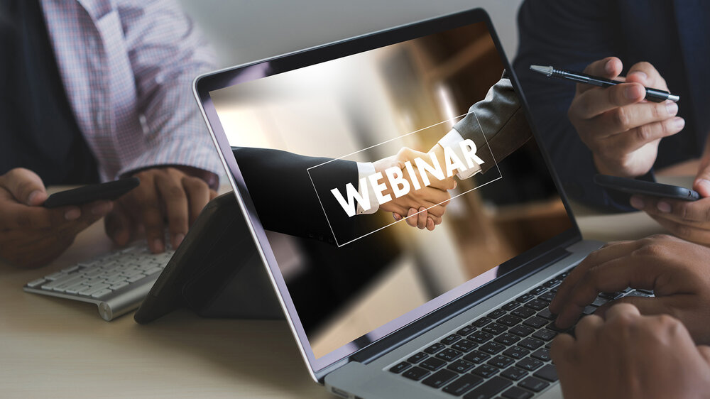 Laptop screen showing a handshake with the word 'WEBINAR' overlaid while two people sit at a desk, one holding a pen and the other a smartphone.