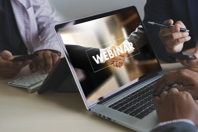 Laptop screen showing a handshake with the word 'WEBINAR' overlaid while two people sit at a desk, one holding a pen and the other a smartphone.