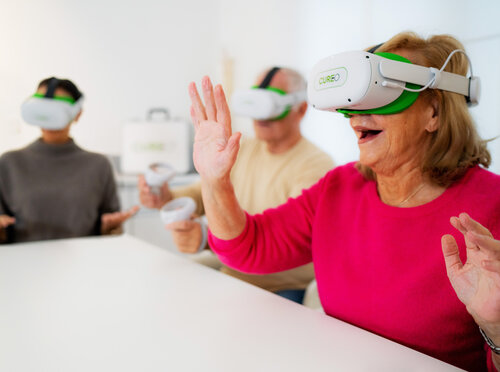 Drei Personen mit Virtual-Reality-Brillen und Controllern, vorne eine Frau im pinken Pullover, die lächelnd ihre Hände gehoben hält.