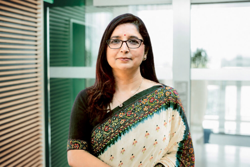 A portrait photograph of a smartly dressed woman standing next to an office desk and leaning on it.