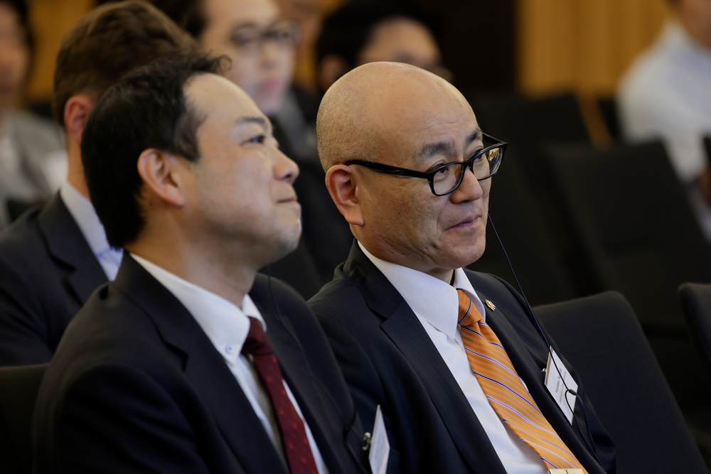 Two men in business suits sit attentively in a conference setting. One wears glasses and an orange tie; the mood is focused and professional.