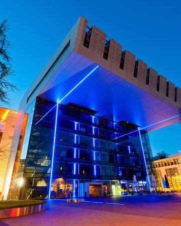 Modernes Gebäude mit blauer Beleuchtung und Glasfassade bei Nacht, umgeben von Bäumen und Straßenbeleuchtung.