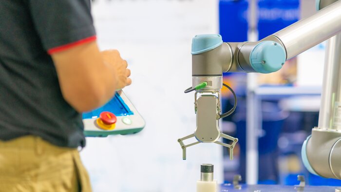 Eine Person steuert mit einem Tablet einen kollaborativen Roboterarm mit Greifer in einer Produktionsumgebung.