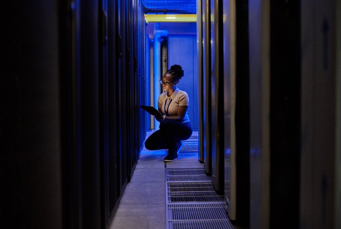 Eine Frau steht mit einem Tablet zwischen Serverracks in einem Rechenzentrum mit blauer Beleuchtung.