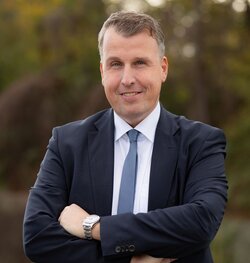 A man in a suit smiles confidently with arms crossed. He wears a watch and a blue tie. Background is a blurred natural setting with trees.