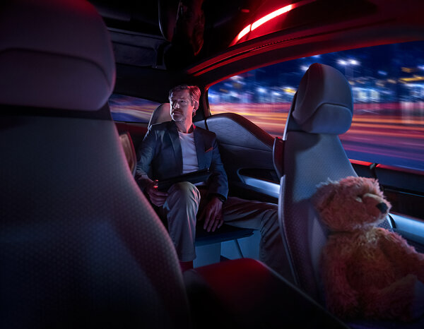 Ein Mann sitzt auf dem Rücksitz eines Autos mit Tablet in der Hand, neben ihm liegt ein Teddybär auf dem Vordersitz, draußen ziehen nachts Lichter vorbei.