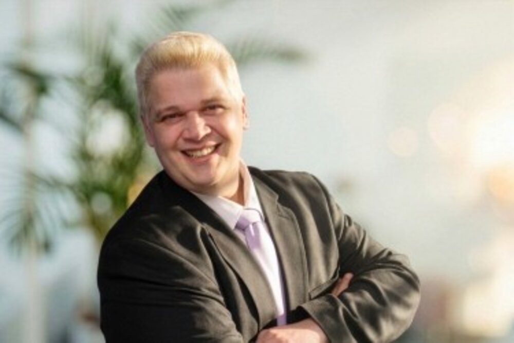 A portrait photograph of a man in a suit with arms crossed standing outdoors with blurred greenery in the background