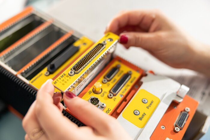 Hands assembling a yellow and orange electronic component with multiple connectors and labels like "Busy" and "Power." The scene conveys precision.