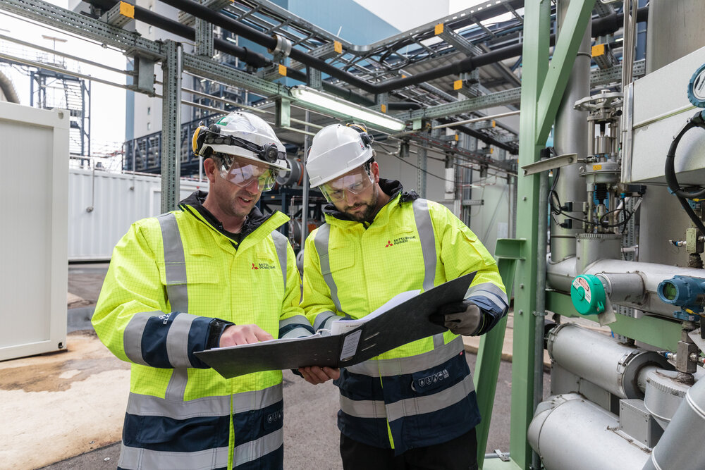 Two men in protective clothing are looking at documents in the middle of an industrial plant with pipework.