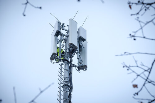 Ein Mobilfunkmast mit mehreren weißen Antennen und Kabeln, ein Techniker in gelber Sicherheitskleidung arbeitet daran.