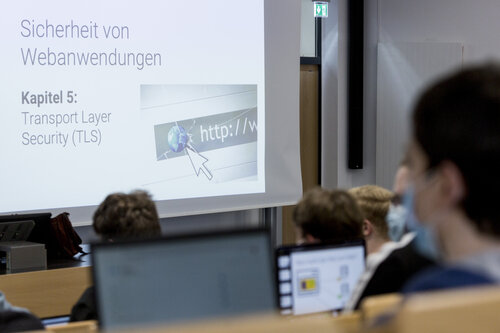 Ein Hörsaal mit Studierenden, unter anderem mit Laptops auf den Tischen. Sie schauen auf eine Präsentationsleinwand.